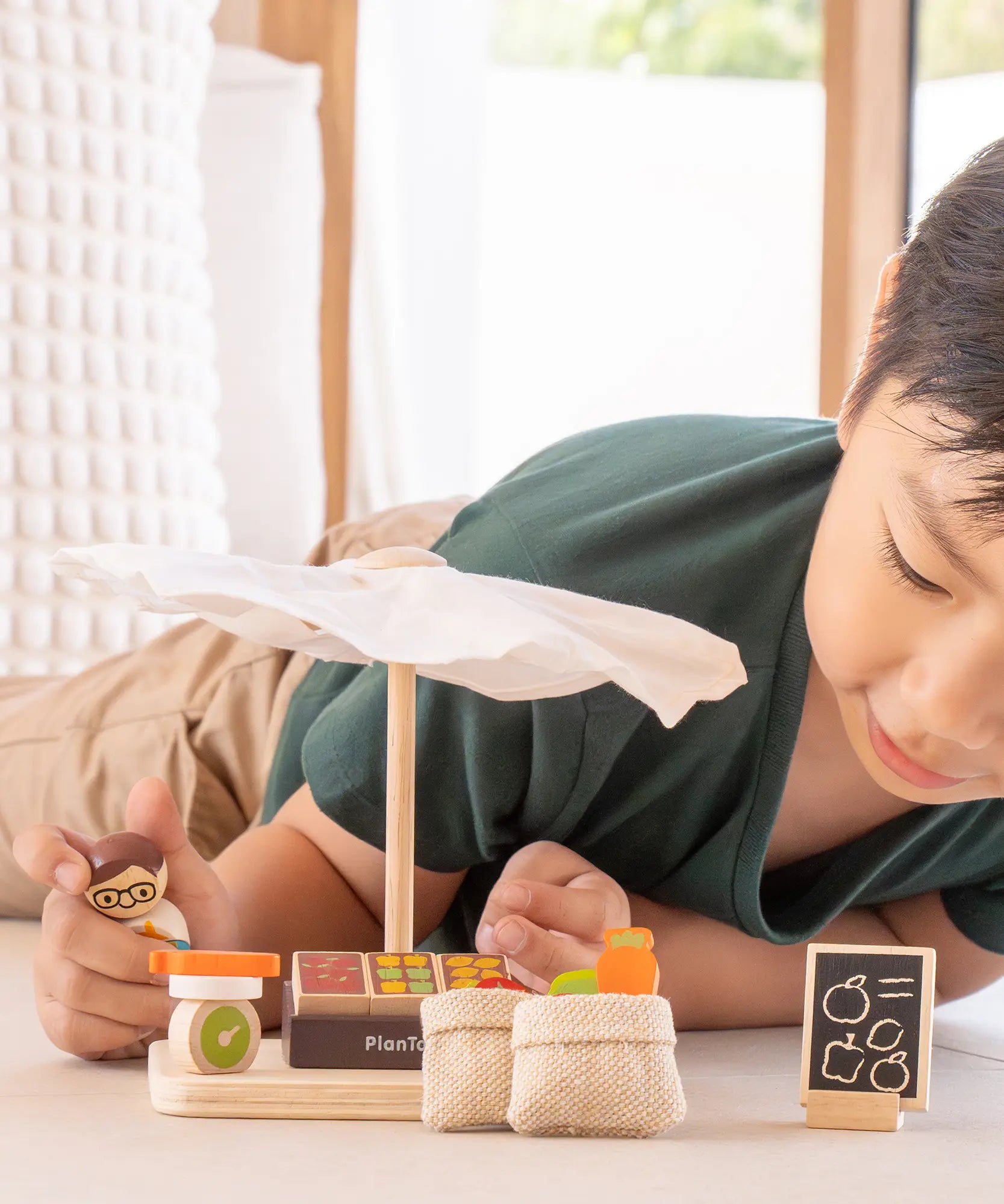 Child playing with the PlanToys Farmer's Market wooden toy playset. The child is placing produce on the wooden toy scales with the worker figure.
