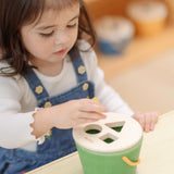 Child placing the yellow block into the green PlanToys geo sorting bucket wooden shape sorting toy. 