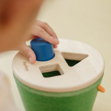 Close up of child placing the blue cylinder into the PlanToys geo sorting bucket wooden shape sorting toy. 