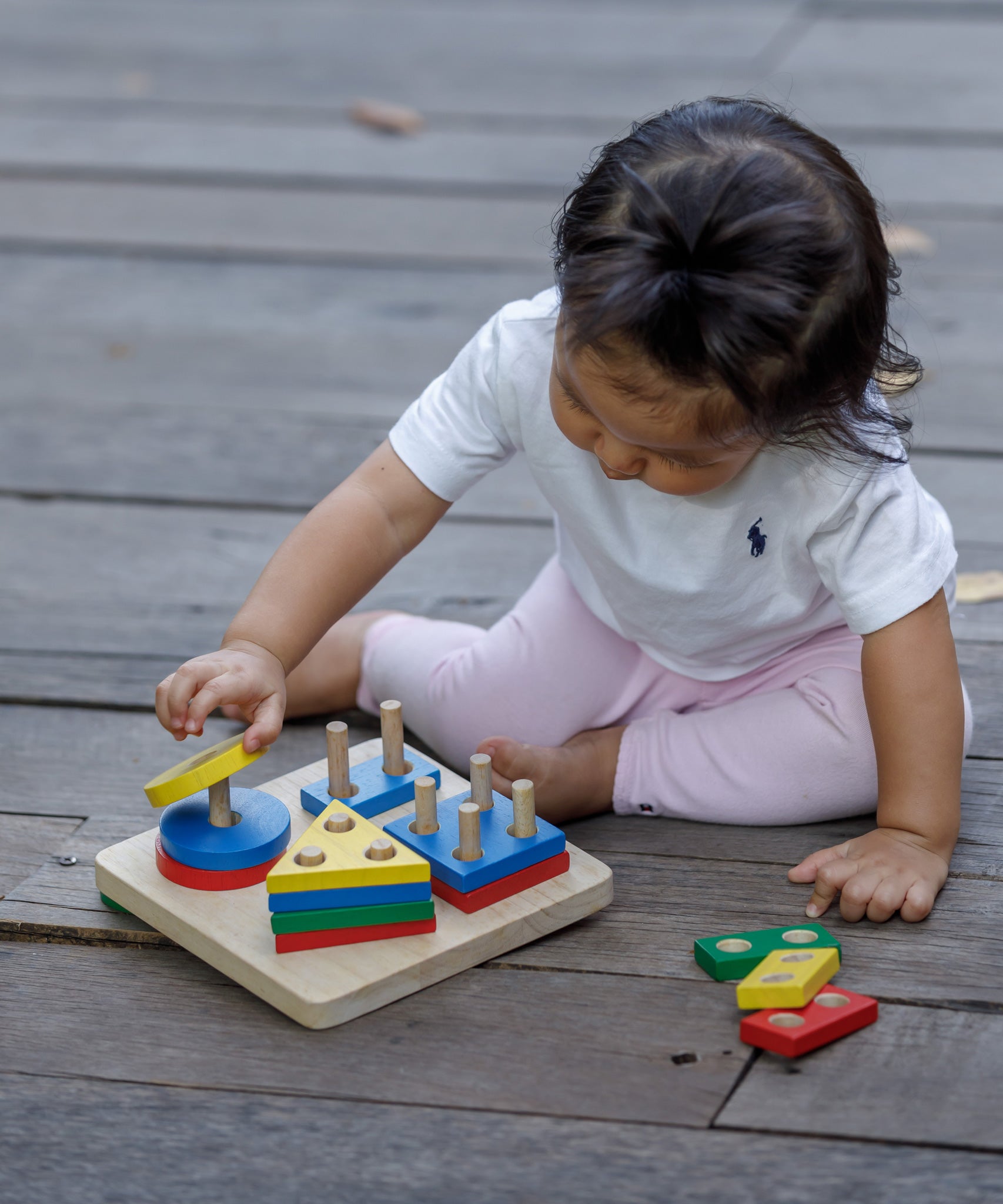 A child sitting on wooden decking playing with the PlanToys Geometric Sorting Board. They are placing the yellow coloured circle piece on top of the other circles. 