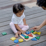 A child sitting on wooden decking playing with the PlanToys Geometric Sorting Board. Many of the pieces in various colours and shapes can be seen taken off the base piece and placed besides the board. 