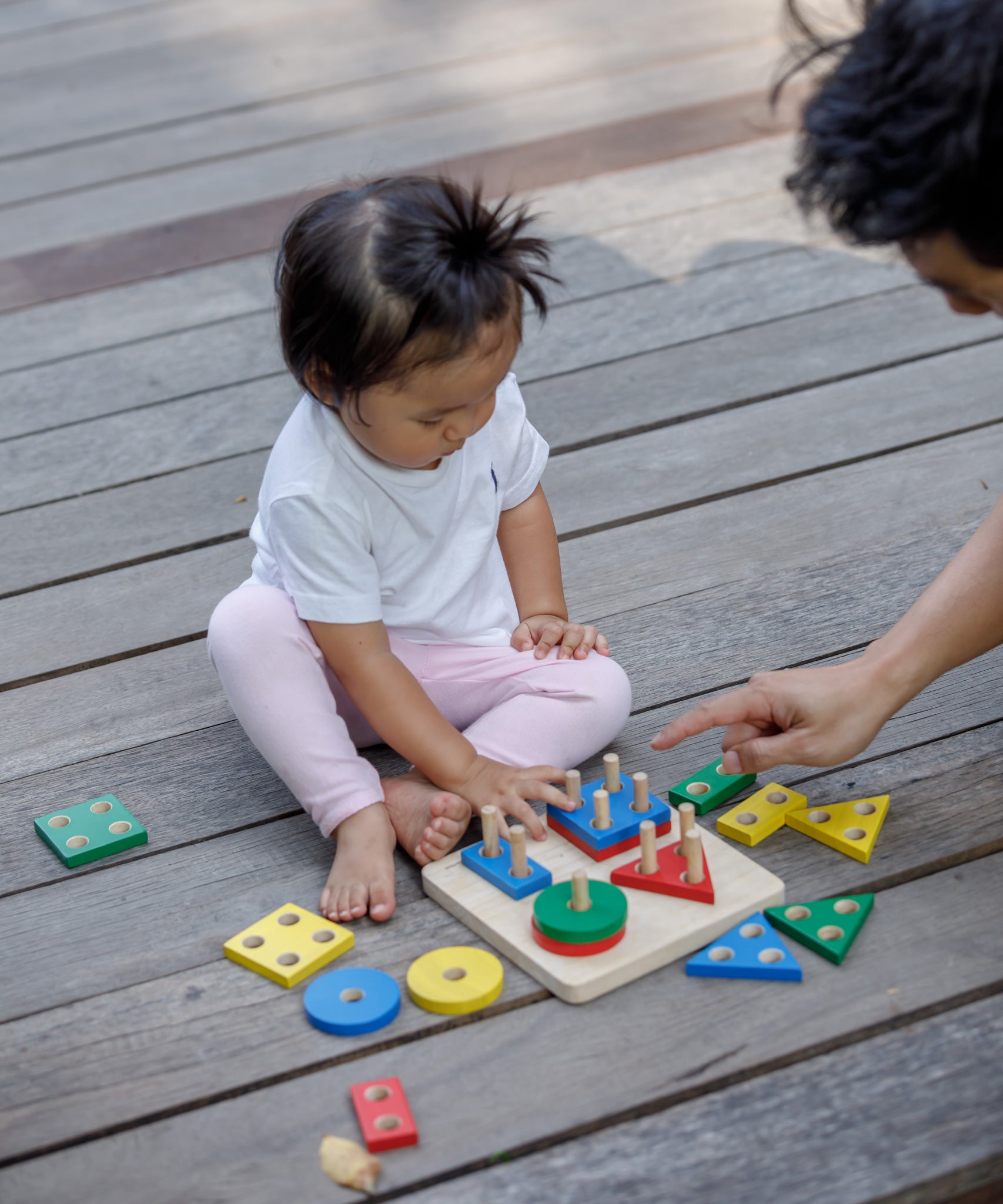 A child sitting on wooden decking playing with the PlanToys Geometric Sorting Board. Many of the pieces in various colours and shapes can be seen taken off the base piece and placed besides the board. 