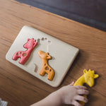 A top view of the PlanToys Giraffe Puzzle on a wooden table. A child is holding the yellow puzzle piece.