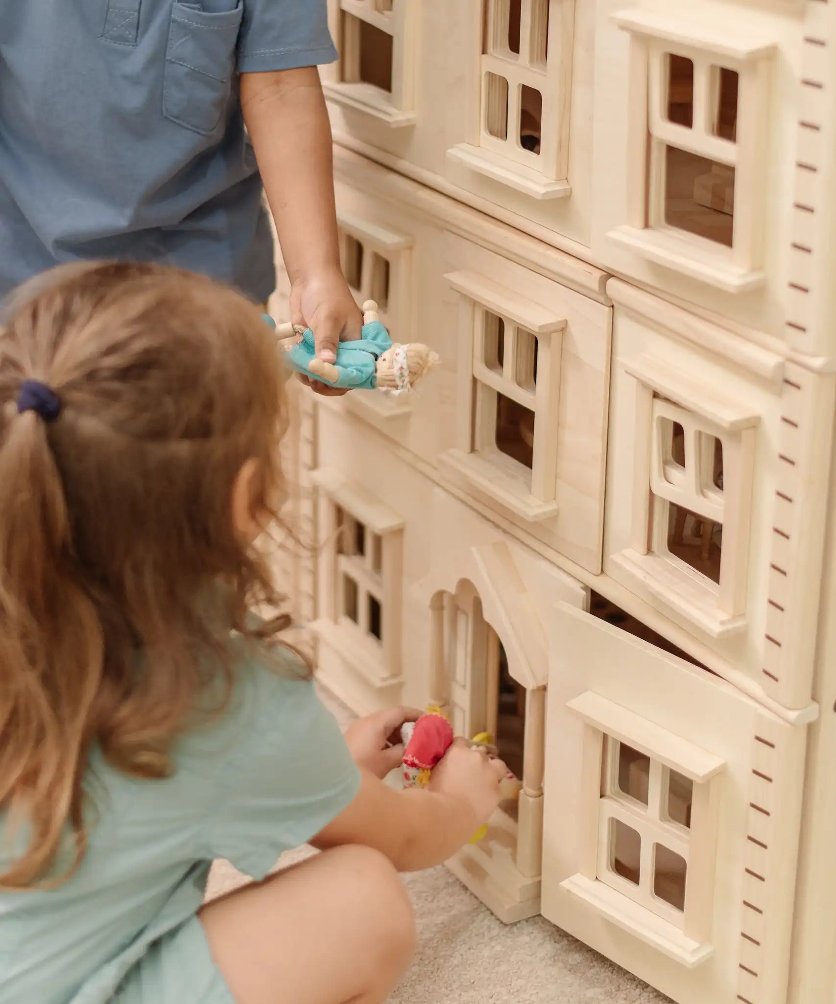 Close up of children playing with the PlanToys Grand Victorian Dollhouse. They are placing doll in the house through the front door. 