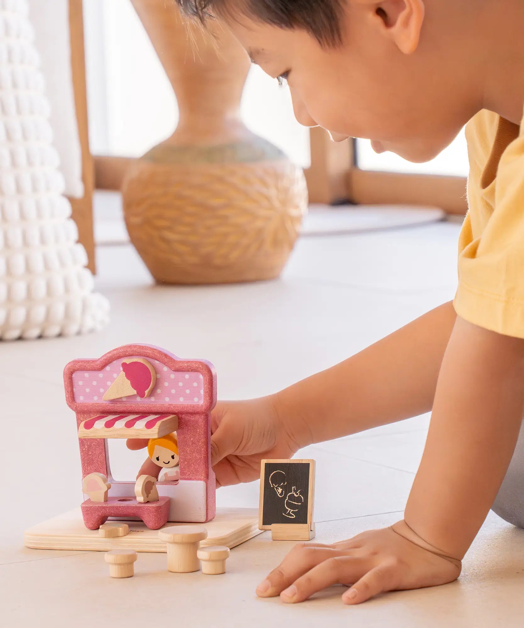 Child playing with the PlanToys ice cream shop wooden toy playset holding the wooden worker figure at the serving window of the shop.