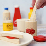Close up of a hand holding a PlanToys kids wooden knife in a wooden pot of PlanToys strawberry jam play food on a grey background