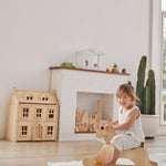 Young girl sat on a PlanToys wooden rocking horse on a white rug in front of a Victorian dollhouse toy