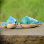 PlanToys Wooden Dolphin Whistle Toy next to the Whale Whistle on a stone wall with grass background.