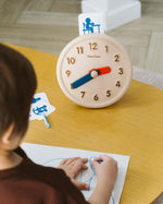 Close up of child drawing on some paper next to the PlanToys wooden activities clock