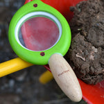 A close up of a Plan Toys Leaf Magnifier toy placed on a side of a red wheelbarrow which is filled with mud. 