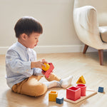 A child sitting on a wooden floor playing with the PlanToys Matching & Nesting Shapes Puzzle.