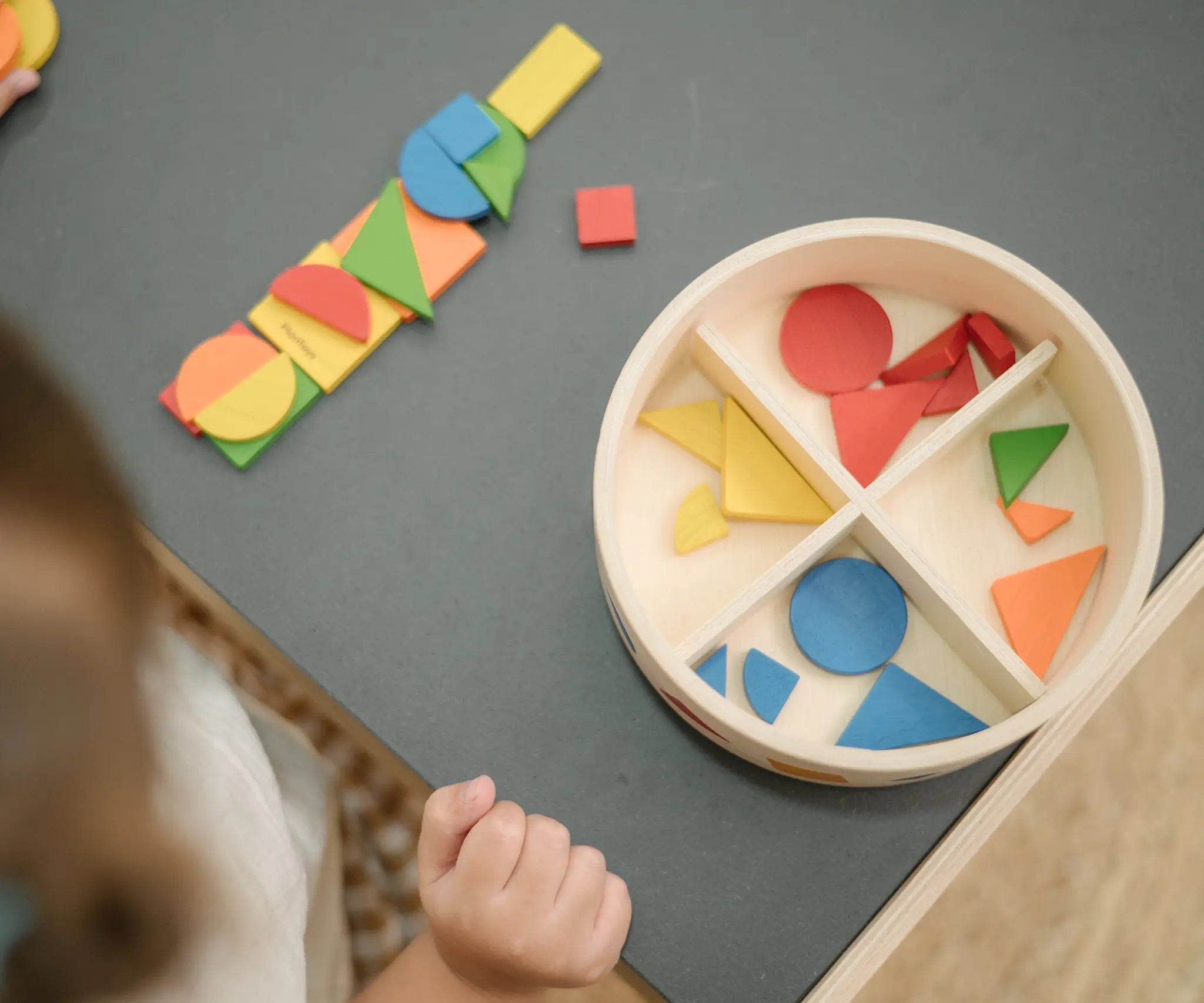 Overhead view of the wooden pieces from the PlanToys Mosaic wooden pattern making toy placed next to the wooden storage box.