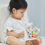 A baby sitting on a bed holding a PlanToys My First Phone toy. 