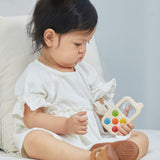 A baby sitting on a bed holding a PlanToys My First Phone toy. 