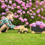 A child playing with the  Plan Toys Special Edition Natural Dump Truck and wooden pull along wagon outdoors on grass. There are purple hydrangeas behind them 
