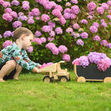 A child playing with the  Plan Toys Special Edition Natural Dump Truck and wooden pull along wagon outdoors on grass. There are purple hydrangeas behind them 