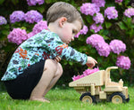 A child playing with the  Plan Toys Special Edition Natural Dump Truck outdoors on grass. There are purple hydrangeas behind them. The child is placing some petals from the hydrangeas into the back of the dumper truck, 