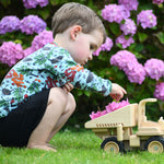 A child playing with the  Plan Toys Special Edition Natural Dump Truck outdoors on grass. There are purple hydrangeas behind them. The child is placing some petals from the hydrangeas into the back of the dumper truck, 