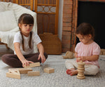 Two children sitting down on a patterned rug, one is playing with the PlanToys Hollow Blocks whilst the other is using the natural PlanToys stacker 