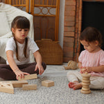 Two children sitting down on a patterned rug, one is playing with the PlanToys Hollow Blocks whilst the other is using the natural PlanToys stacker 