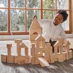 A child playing with the Plan Toys natural wooden 50 Unit Building Block set. The child has built a long building with multiple turret like pillars and a tall tower in the middle. The child can be seen sitting behind the structure. 
