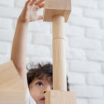 A close up of a child playing with the Plan Toys natural wooden 50 Unit Building Block set. the child can be seen looking up at a pile of clocks reaching for the square shaped block that has been stacked on top of cylinder shaped blocks. 