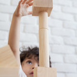 A close up of a child playing with the Plan Toys natural wooden 50 Unit Building Block set. the child can be seen looking up at a pile of clocks reaching for the square shaped block that has been stacked on top of cylinder shaped blocks. 