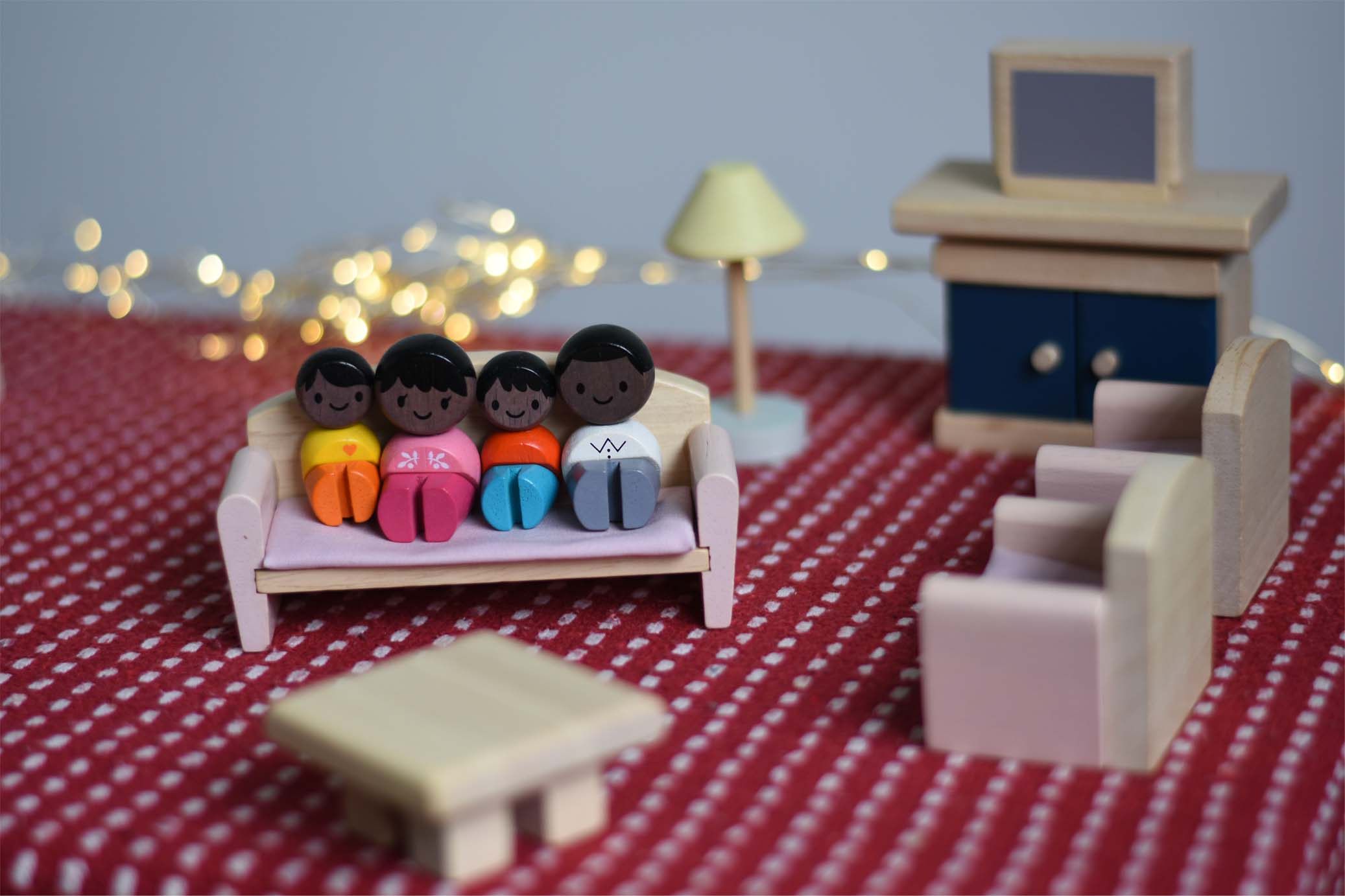A family of PlanToys peg dolls sitting on the wooden dollhouse sofa amongst other living room furniture. 