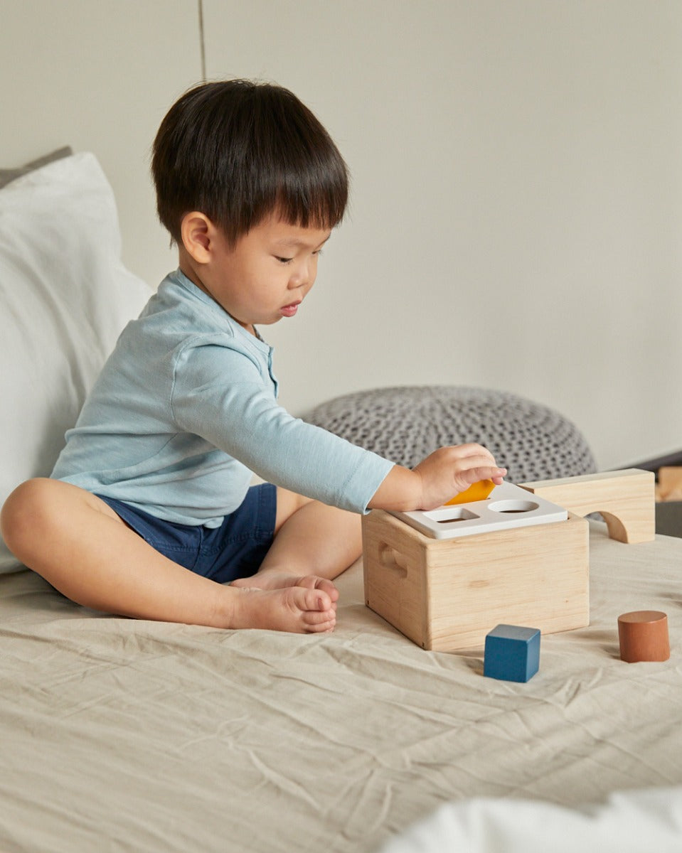 Child sat on a bed putting wooden shape blocks into the PlanToys wooden shape sorter box