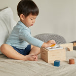 Child sat on a bed putting wooden shape blocks into the PlanToys wooden shape sorter box
