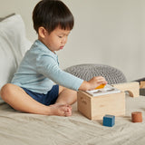 Child sat on a bed putting wooden shape blocks into the PlanToys wooden shape sorter box