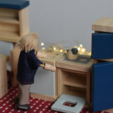 A PlanToys peg doll positioned in a small-world play scene, using the wooden dollhouse kitchen furniture.