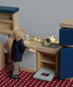 A PlanToys peg doll positioned in a small-world play scene, using the wooden dollhouse kitchen furniture.