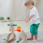 A child walking towards the PlanToys Palomino Rocking Horse. The rocking horse is on a grey coloured rug. 
