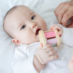 A baby is laying down on a white sheet holding a PlanToys Pastel Roller up to their mouth. 
