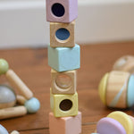 The PlanToys Pastel Sensory Blocks stacked up on a wooden floor. Other PlanToys pastel baby toys can be seen around them.