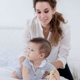 A bay playing with a PlanToys Pastel Triangle Clutching Toy on a white bed, an adult is holding onto them. 