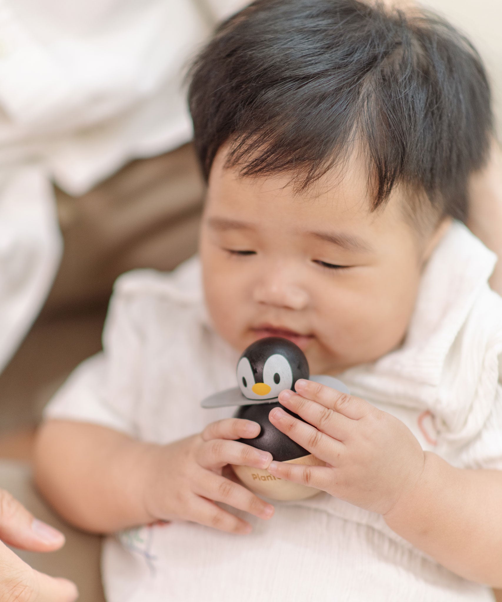 A baby holding the PlanToys Penguin Wobbler wooden baby toy. A circular shaped penguin toy that wobbles when placed on a flat surface. PlanToys range of toys are made from sustainably sourced rubberwood, we're proud stockists of PlanToys here at Babipur. 