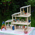 The living room, kitchen / diner, bathroom and bedroom Plan Toys PlanSpace sets shown stacked on top of each other to create a 3 storey house. The levels can be arranged to the users preference offering a fully customised set up. Various wooden peg dolls and small wooden loose parts have been dotted around the house such as small wooden pizzas on the dining table.
