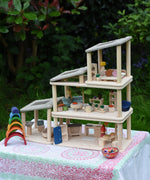 The living room, kitchen / diner, bathroom and bedroom Plan Toys PlanSpace sets shown stacked on top of each other to create a 3 storey house. The levels can be arranged to the users preference offering a fully customised set up. Various wooden peg dolls and small wooden loose parts have been dotted around the house such as small wooden pizzas on the dining table.