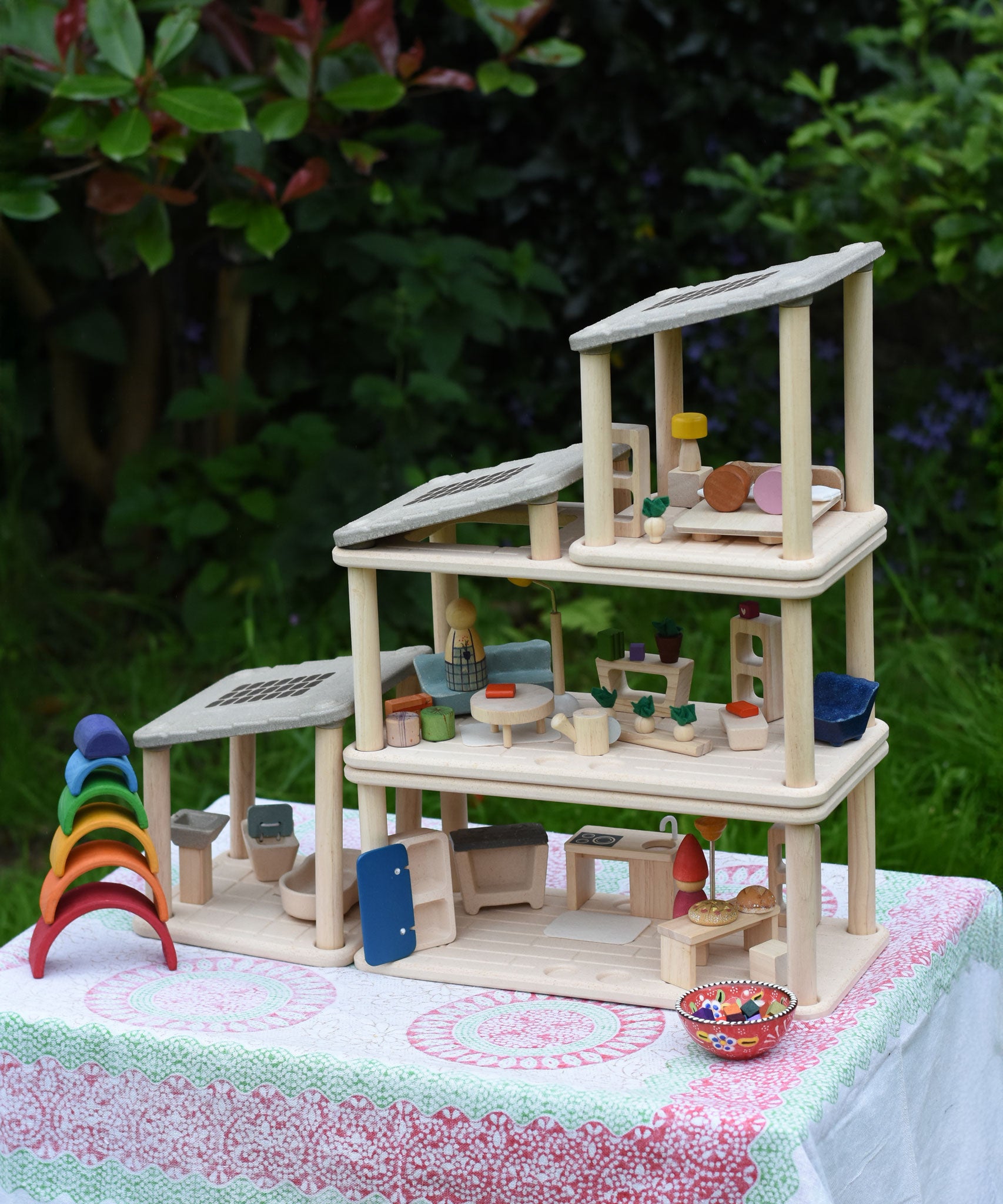 The living room, kitchen / diner, bathroom and bedroom Plan Toys PlanSpace sets shown stacked on top of each other to create a 3 storey house. The levels can be arranged to the users preference offering a fully customised set up. Various wooden peg dolls and small wooden loose parts have been dotted around the house such as small wooden pizzas on the dining table.