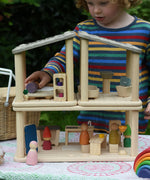 A child playing with the PlanToys PlaySpace dolls house. The kitchen diner, bedroom and bathroom sets have been stacked up to create a two storey house. There are wooden peg dolls placed in various locations around the house.