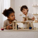 2 children playing the with Plantoys wooden planspace living room set on a concrete surface showing the sunlight piece being out on top of the living room