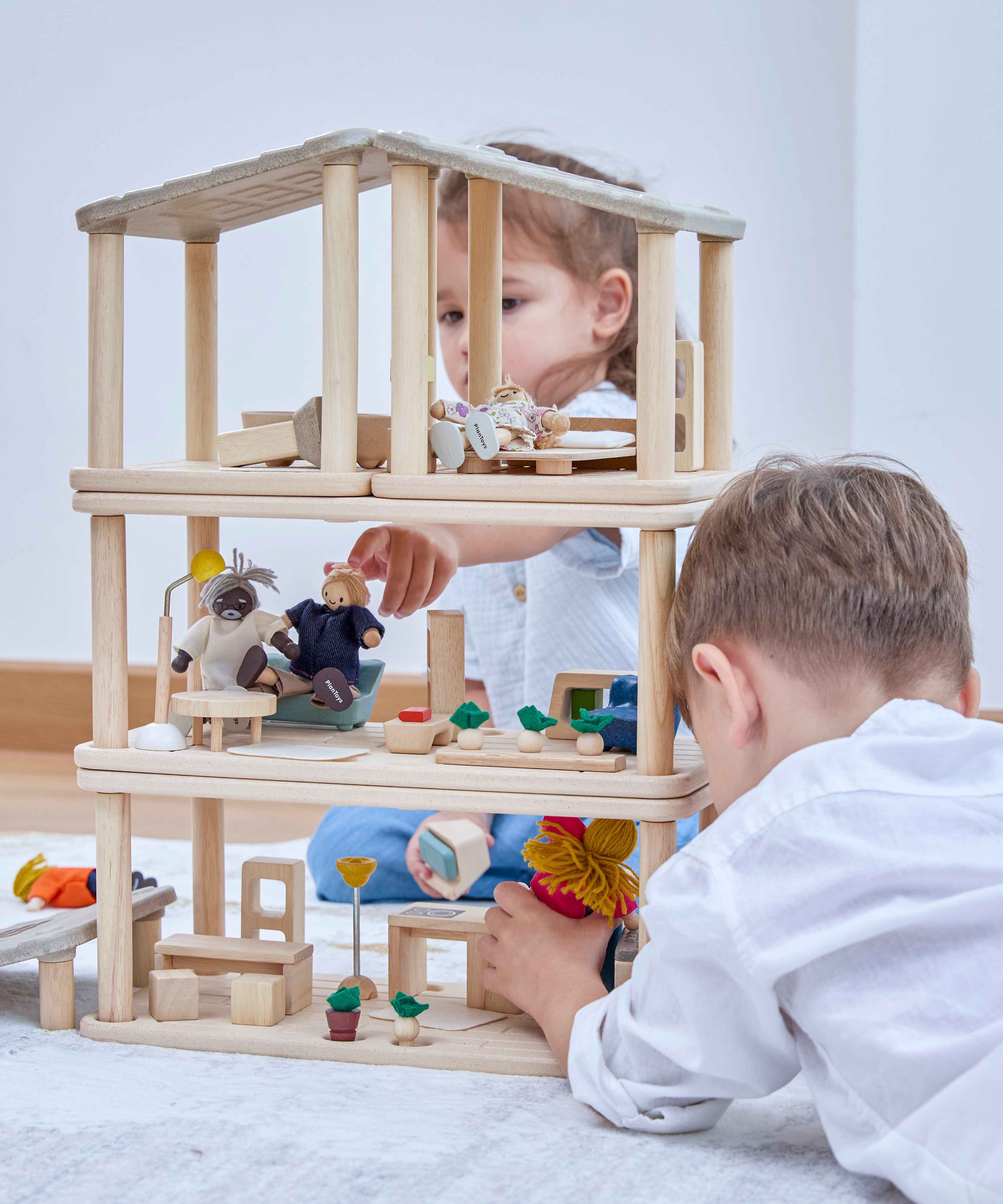 2 children playing playing the with Plantoys wooden planspace set that's stacked on top of eachother to create a dollhouse, showing all of the accessories and some of the PlanToys family dolls 
