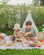 Two children sat on a blanket in front of a small tent playing with wooden play food on top of the PlanToys wooden BBQ set