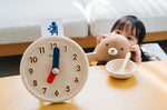 Young child crouched down holding a teddy bear next to an empty bowl beside the PlanToys wooden activity clock