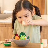 Child holding a wooden pepper shaker over the PlanToys chef salad bowl full of loose play food