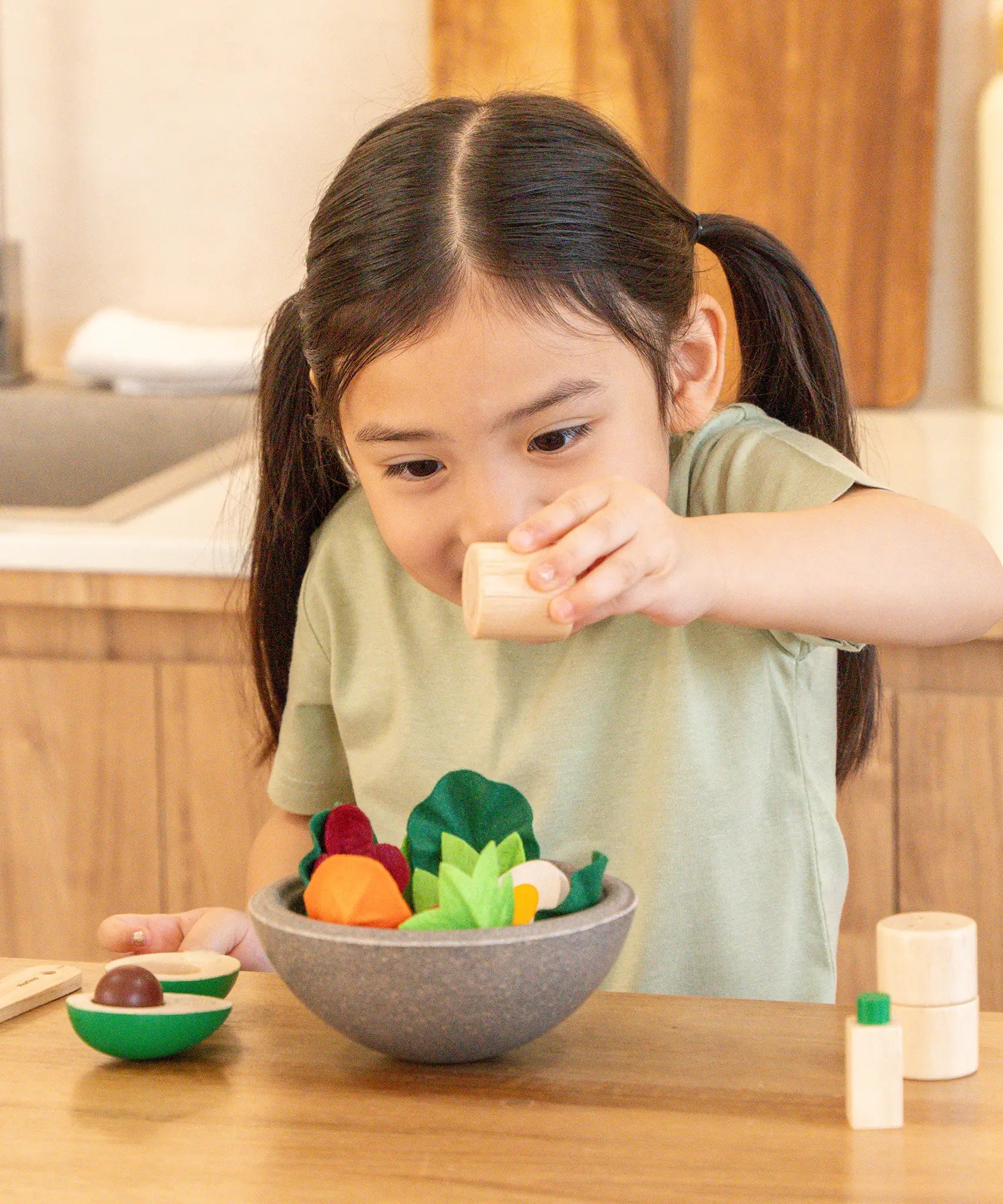 Child holding a wooden pepper shaker over the PlanToys chef salad bowl full of loose play food