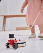 Close up of young girl in a pink dress pulling a PlanToys musical pull along drummer toy on a white floor