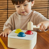 Toddler playing with the PlanToys punch and drop bin wooden pounding toy. Hammering the red ball into the bin through a slot on the lid.